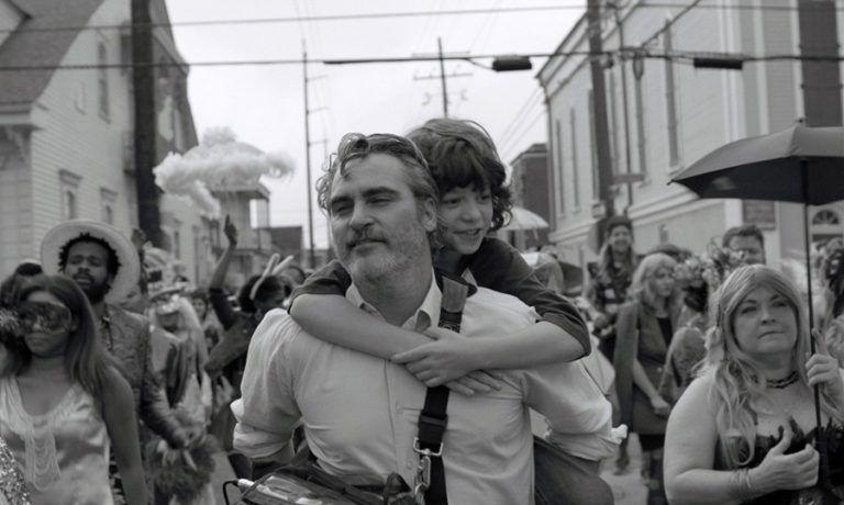 Kadr z filmu. Mężczyzna z chłopcem na ramionach. Wokół tłum ludzi w małym miasteczku. Fotografia czarnobiała.